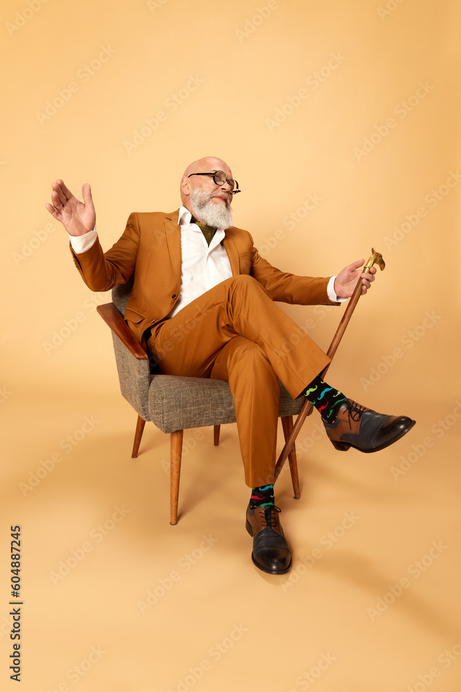 Fototapeta premium Portrait of bearded, mature, bald man in elegant, classical suit and walking stick, sitting in armchair and smiling against studio background. Concept of human emotions, lifestyle, fashion, business