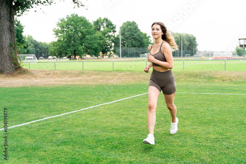 Young blond slim fit sporty woman with well-groomed long hair in stylish fitness clothes and sneakers jogging outside on football grass lawn field on summer day. Fitness, jogging, footwear, lifestyle