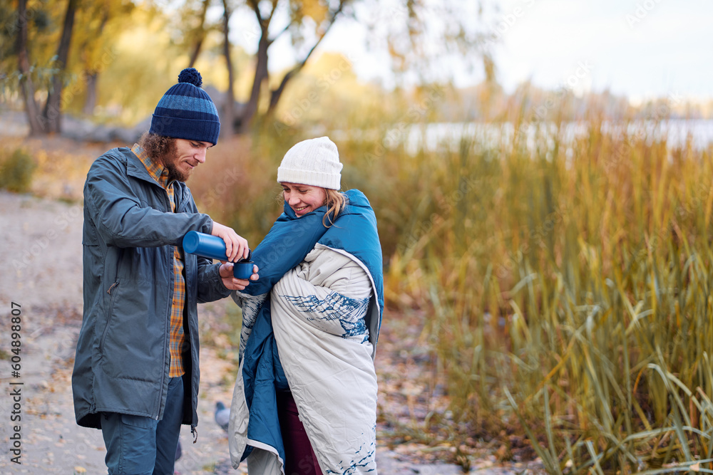 Fototapeta premium Holidays with friend. Young couple drink hot beverage from thermos near river.