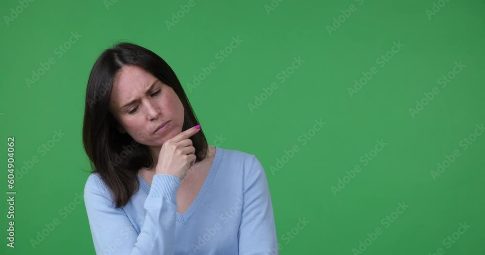 Caucasian woman stands on a green chroma key, looking pensive and lost in thought. She places her hand on her chin and contemplates her options, considering her decision carefully.