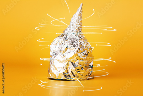 Hat made of aluminium foil on yellow background. Afraid of 5G radiation or aliens. Symbol for conspiracy theory