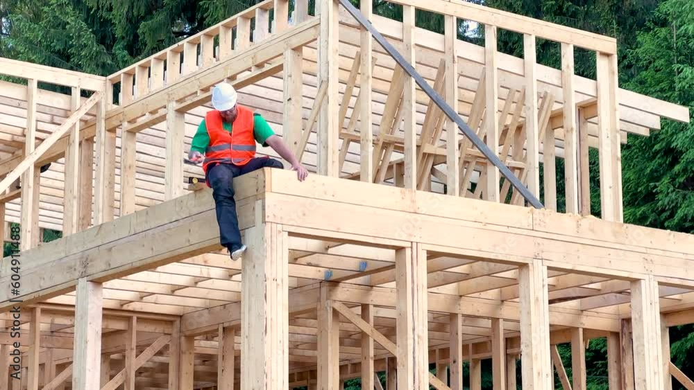 Carpenter constructing two-story wooden frame house. Bearded man in glasses hammering nails into structure while wearing protective helmet, construction vest. Concept of modern ecological construction Carpenter constructing two-story wooden frame house. Bearded man in glasses hammering nails into structure while wearing protective helmet, construction vest. Concept of modern ecological construction