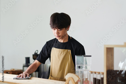 Fotografie Pensive young Asian barista cleaning counter before opening cafe in the morning