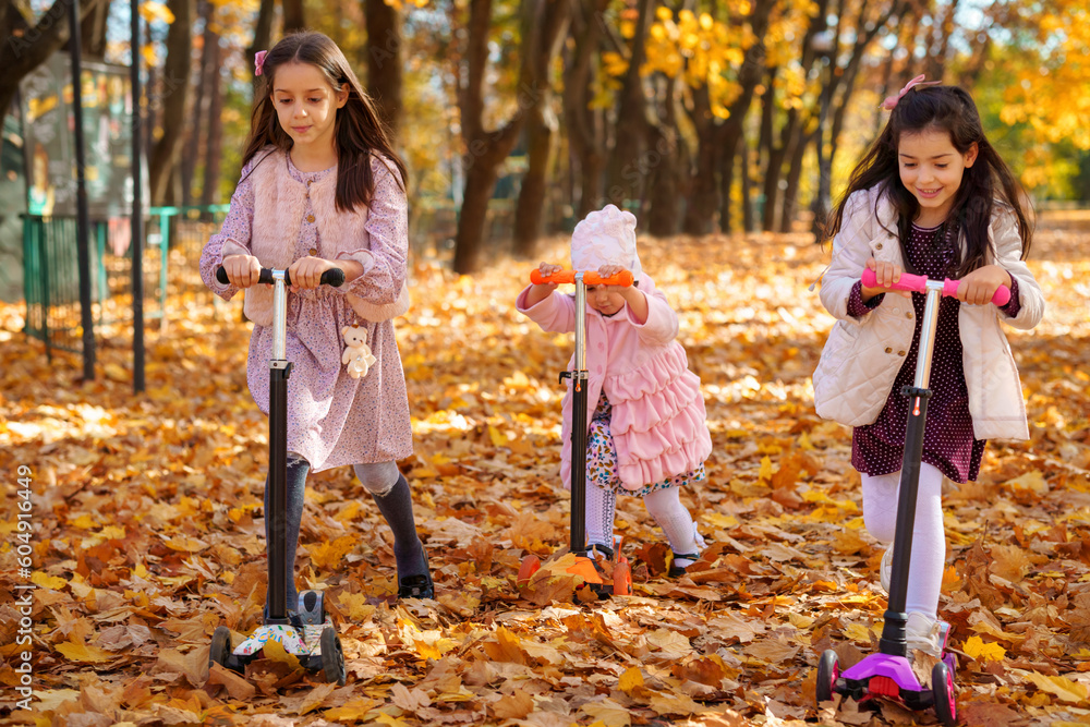 sister girls ride scooters in the autumn city park, they play, have fun and smile, yellow maple leaves around, beautiful nature, bright sunny day sister girls ride scooters in the autumn city park, they play, have fun and smile, yellow maple leaves around, beautiful nature, bright sunny day