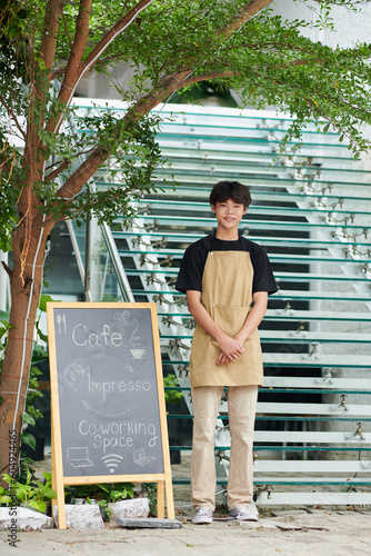 Positive coffeeshop barista on apron standing next to signboard photo