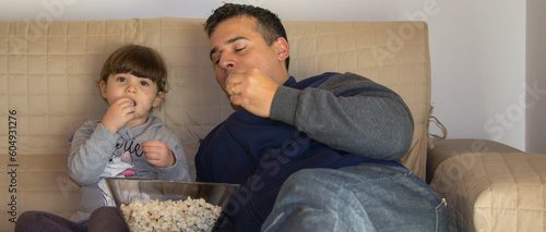 Image of a dad with his daughter lying on the sofa at home while watching TV and eating popcorn. Horizontal banner 