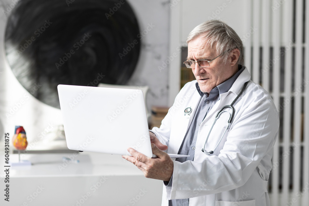 Senior doctor stands at the desktop and works on laptop in his modern medical office. Online consultation concept Senior doctor stands at the desktop and works on laptop in his modern medical office. Online consultation concept