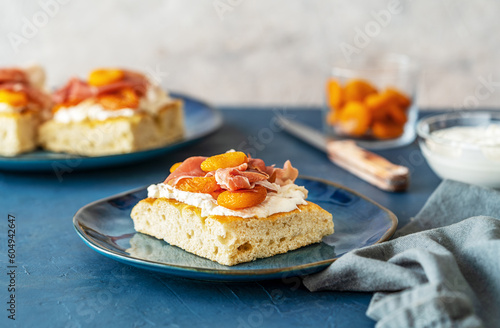 Focaccia with prosciutto, soft cheese stracciatella, dry apricots and pistachios on a blue plate, ingredients, others sandwiches backside over dark blue and grey background