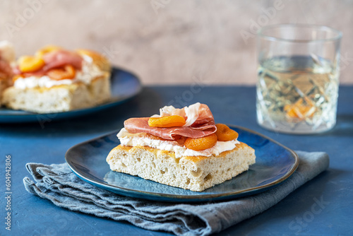 Focaccia with prosciutto, soft cheese buratta, dry apricots on a blue plate, grey napkin, fresh drink and others sandwiches backside over dark blue and grey background