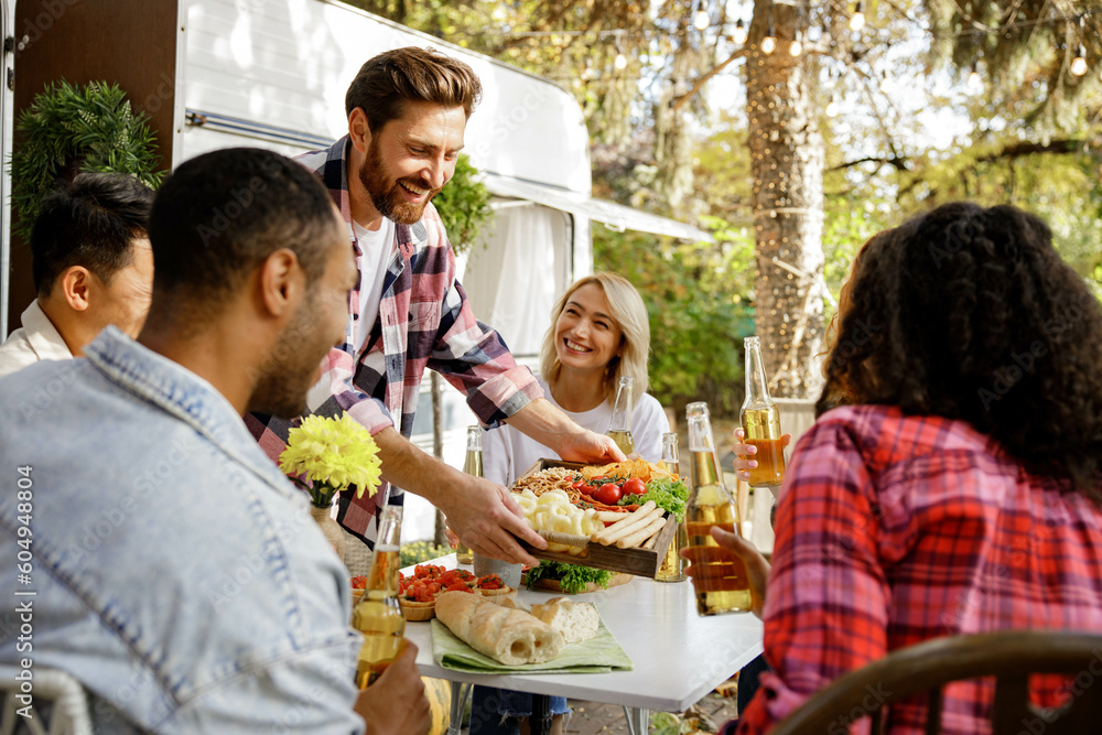 A cheerful multinational company is having fun on a picnic. Multisex friends sitting at the table outdoors celebrating eating and drinking drinks. A cheerful multinational company is having fun on a picnic. Multisex friends sitting at the table outdoors celebrating eating and drinking drinks.