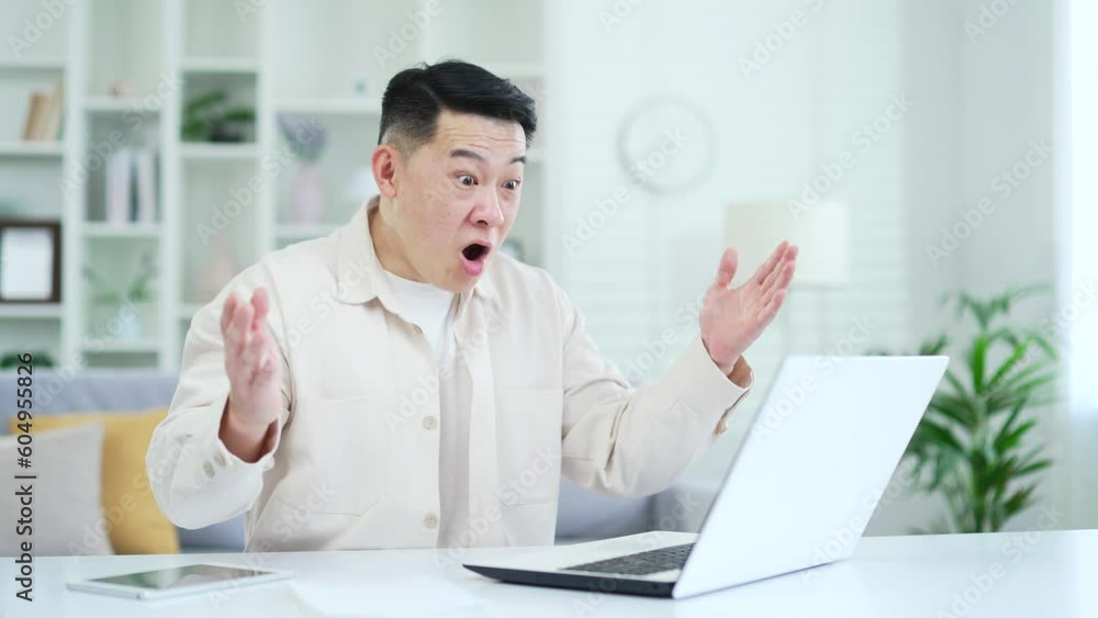 Happy asian man celebrating business success reading great news on laptop while sitting at desk at workplace in home office. Excited male is satisfied with a pleasant notification, shows a yes sign Happy asian man celebrating business success reading great news on laptop while sitting at desk at workplace in home office. Excited male is satisfied with a pleasant notification, shows a yes sign