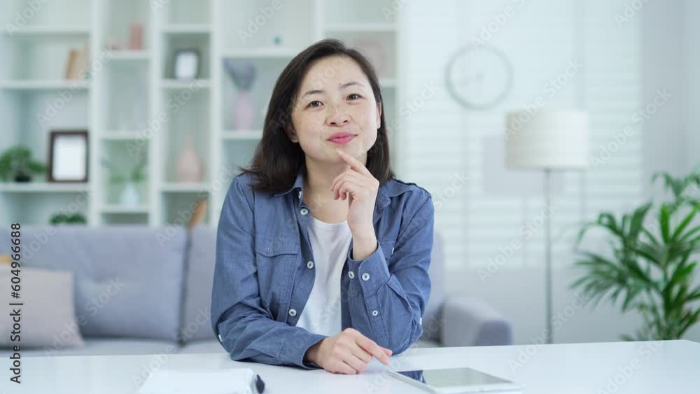 Webcam view. Asian woman watching video call lesson or training looking at camera while sitting in home office. Female is studying online, listening remotely to a course, webinar or teacher's lecture Webcam view. Asian woman watching video call lesson or training looking at camera while sitting in home office. Female is studying online, listening remotely to a course, webinar or teacher's lecture
