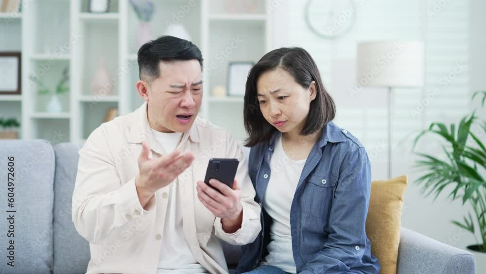 Disappointed asian couple reading bad news on smartphone while sitting on sofa in living room at home. A sad husband and wife are depressed by an unpleasant message, shocked by information about loss Disappointed asian couple reading bad news on smartphone while sitting on sofa in living room at home. A sad husband and wife are depressed by an unpleasant message, shocked by information about loss
