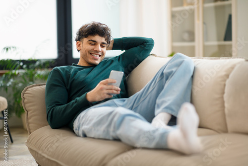 Guy Browsing Internet On Smartphone Lying On Couch At Home