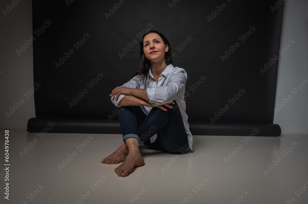 business portrait of a young girl about 40 years old, cute, emotional thoughtful, sitting on the floor on a dark background, indoor, reflected light, shot with copy space