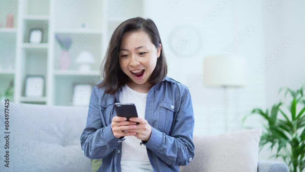 Happy asian woman celebrating success by reading great news on smartphone while sitting on sofa in living room at home. Excited smiling businesswoman satisfied with pleasant message, shows yes gesture Happy asian woman celebrating success by reading great news on smartphone while sitting on sofa in living room at home. Excited smiling businesswoman satisfied with pleasant message, shows yes gesture