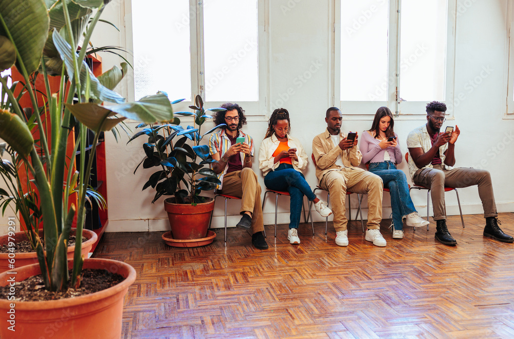 Five multiracial people using cell phones while sitting by wall.