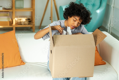 African girl unpacking delivery looking in box. Happy woman opening carton box. Female getting parcel looking at delivered goods items. Satisfied client positive feedback quick express deliver