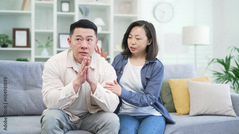 Asian wife tries to reconcile with the offended husband while sitting on the sofa in the living room at home. A woman hugs a man, forgiveness. The couple is talking. The concept of family relations Asian wife tries to reconcile with the offended husband while sitting on the sofa in the living room at home. A woman hugs a man, forgiveness. The couple is talking. The concept of family relations