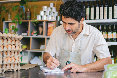 Slika na platnu middle-aged latin man taking inventory in his organic store, writing on a paper