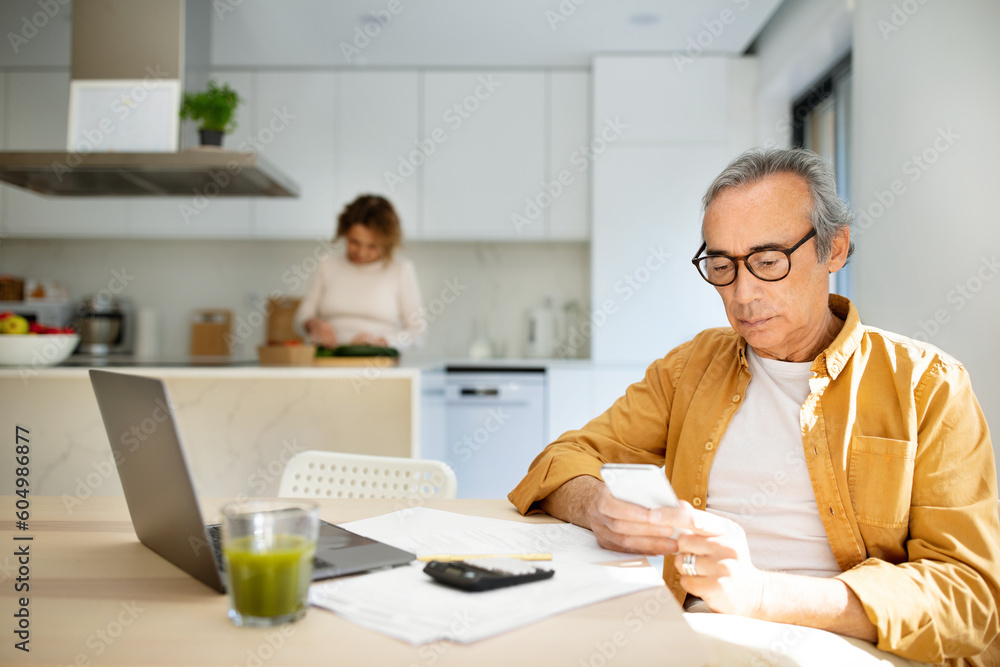 Serious aged man using cellphone, sitting at table in front of laptop in kitchen while his wife cooking on background Serious aged man using cellphone, sitting at table in front of laptop in kitchen while his wife cooking on background