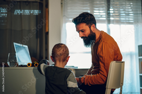 Obraz na płótnie A little boy is standing next to his working father and bothering him during his father's remote job time at the home office