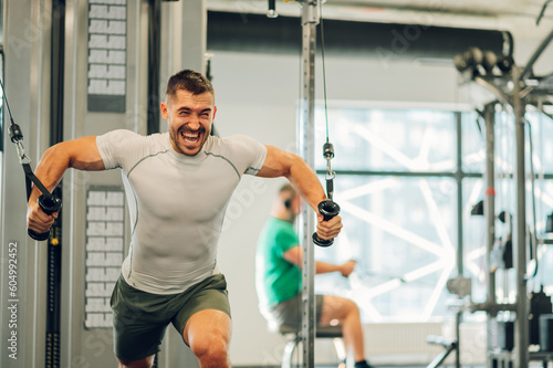 A sportsman is shouting in a gym while lifting heavy weights on a cable machine in a gym.