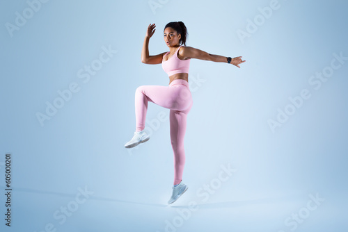Fototapeta Naklejka Na Ścianę i Meble -  Young black woman in sportswear jogging and jumping looking at copy space, sprinting on blue background, full length
