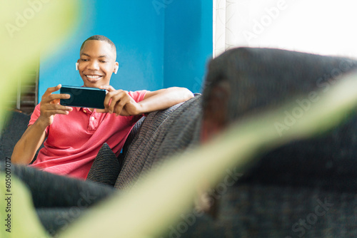 Billede på lærred A happy black african man listens and watches the content of the phone sitting on a sofa in the day at home