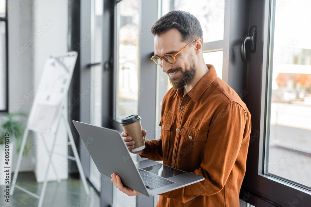 Fototapeta premium brunette and bearded businessman in eyeglasses and stylish shirt holding disposable cup with coffee and looking at laptop near flip chart on blurred background in office