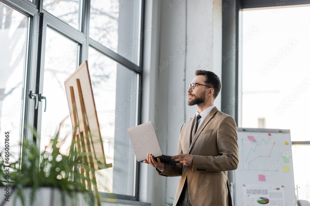Fototapeta premium confident and fashionable bearded businessman with laptop looking away through windows near flip charts with infographics and blurred plants in contemporary office