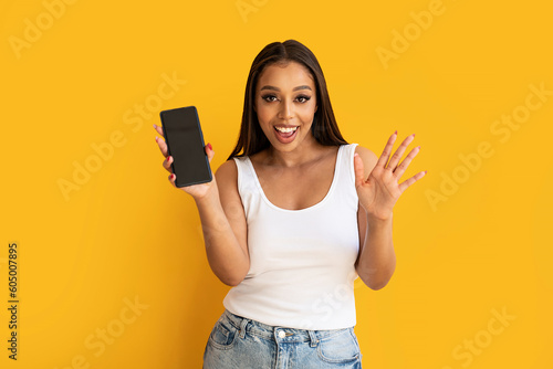 Young happy woman wearing basic clothes smiling to the camera and showing cellphone screen, posing over yellow studio background.