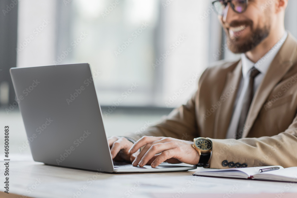 cropped view of smiling and successful bearded businessman in beige blazer and stylish eyeglasses chatting on laptop near notebook and pen on office desk cropped view of smiling and successful bearded businessman in beige blazer and stylish eyeglasses chatting on laptop near notebook and pen on office desk