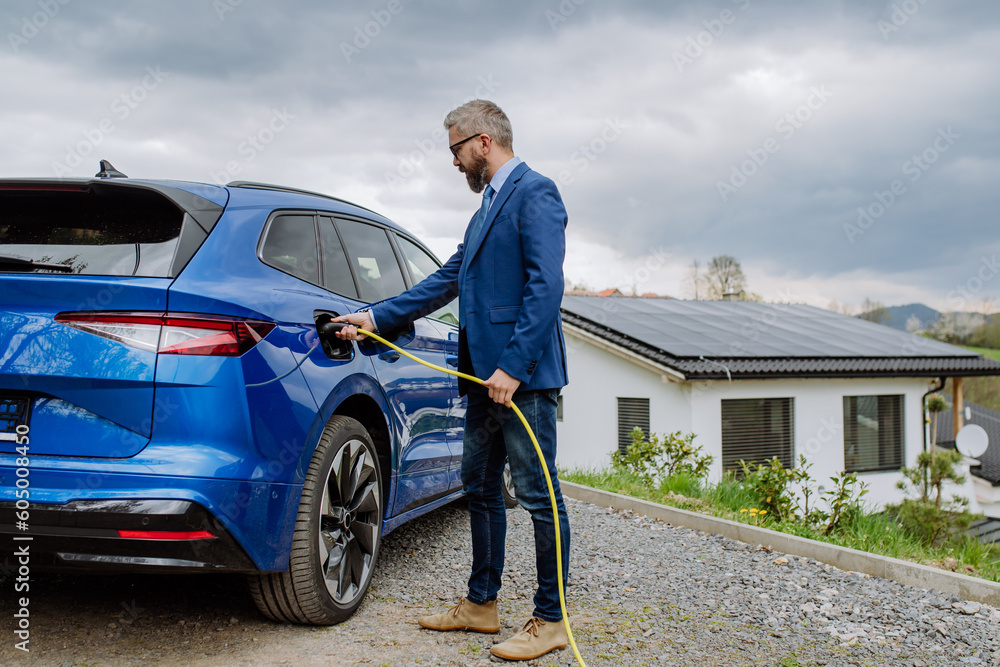 Mature businessman holding power supply cable and charging his electric car. Mature businessman holding power supply cable and charging his electric car.