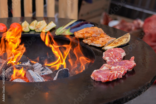 Process of cooking meat steaks, zucchini, eggplant, tomato on grill, brazier at outdoor summer local food market - close up. Gastronomy, cookery and street food concept photo