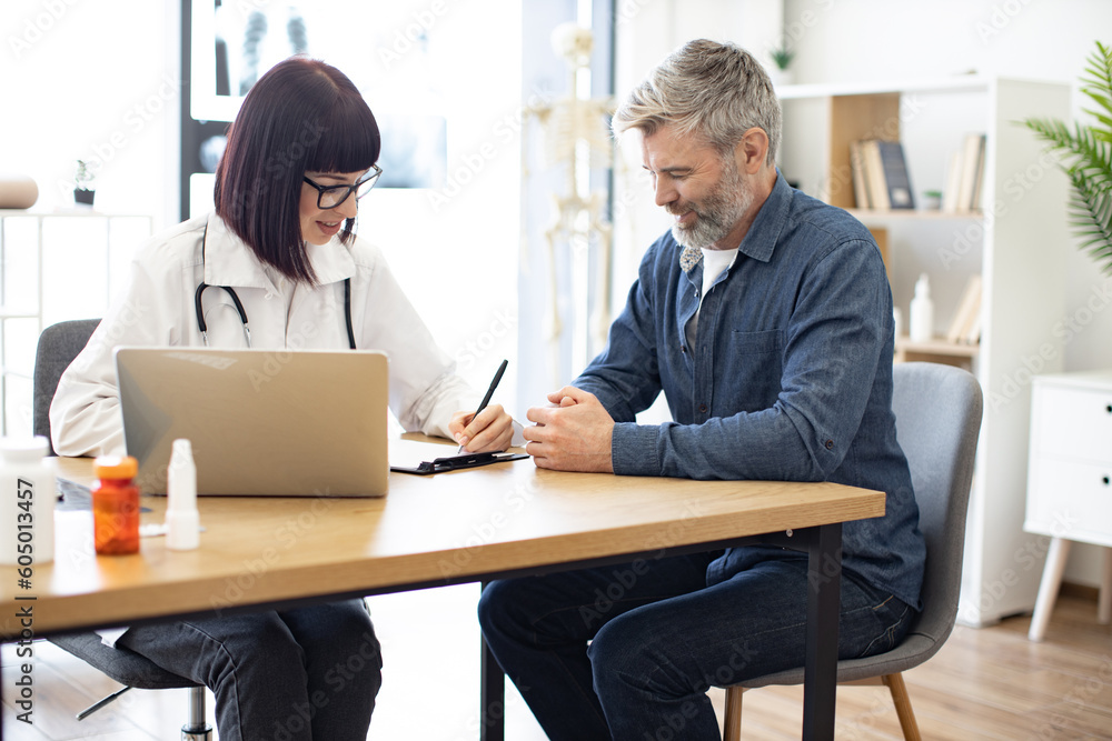 Silver-bearded gentleman describing symptoms of illness to female doctor and gesturing hands. Dark haired physician giving recommendations and using laptop for writing electronic prescription. Silver-bearded gentleman describing symptoms of illness to female doctor and gesturing hands. Dark haired physician giving recommendations and using laptop for writing electronic prescription.