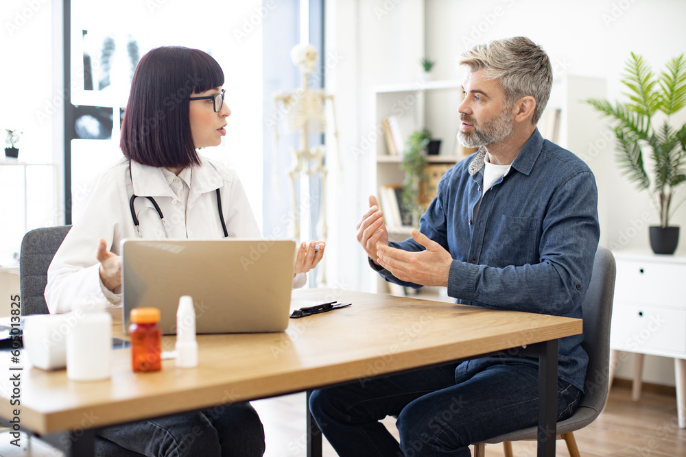 Silver-bearded gentleman describing symptoms of illness to female doctor and gesturing hands. Dark haired physician giving recommendations and using laptop for writing electronic prescription. Silver-bearded gentleman describing symptoms of illness to female doctor and gesturing hands. Dark haired physician giving recommendations and using laptop for writing electronic prescription.