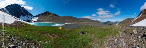 Back country camping area in northern arctic Canada during summer time with panoramic view over glacial lake, yellow tent, huge mountains, snow, ice, green grass. 