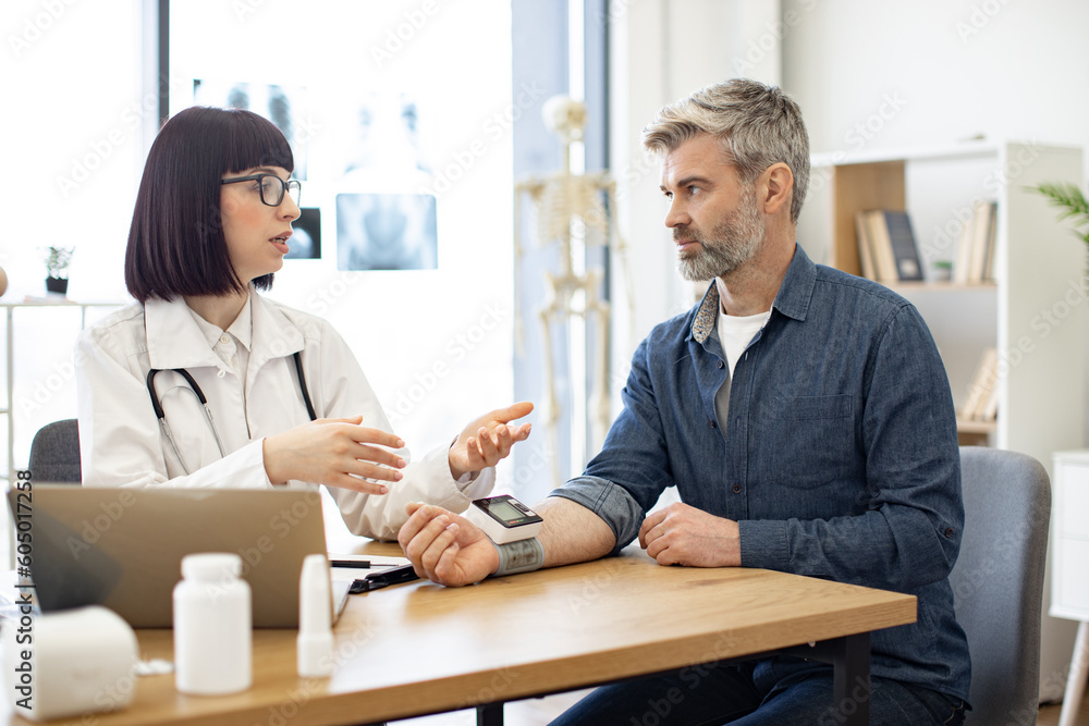 Obraz premium Attractive cardiologist telling patient about all possible reasons of hypertension and keeping medical history on laptop. Adult man listening explanations about necessity of leading healthy lifestyle.