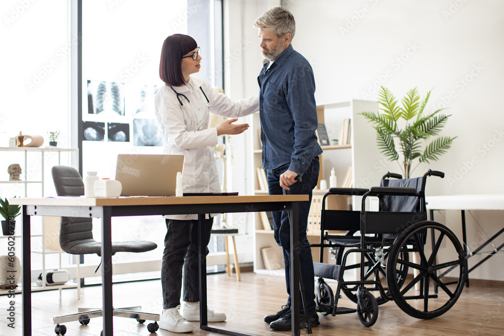 Full length view of man standing near wheelchair with cane while physician giving him hand in doctor's workplace. Medical person encouraging patient after successful treatment in medical center. Full length view of man standing near wheelchair with cane while physician giving him hand in doctor's workplace. Medical person encouraging patient after successful treatment in medical center.