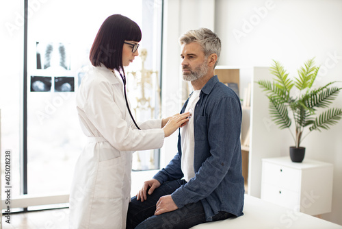 Side view of focused lady in white coat using stethoscope while mature man in casual clothes sitting on exam couch. General practitioner examining heartbeat while providing regular checkup in clinic. © sofiko14