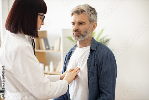 Close up view of serious caucasian man in everyday wear visiting family doctor for full examination in general practice. Young woman in glasses and lab coat checking lungs and heart via stethoscope. photo