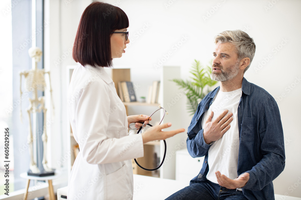 Obraz premium Worried gray-haired man sitting on exam couch with hand on chest while mindful woman in doctor's coat holding stethoscope. Male patient describing symptoms to medical specialist in consulting room.