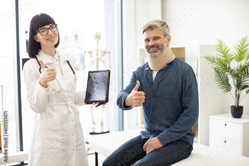 Cheerful young woman with digital tablet and smiling mature man in cervical collar posing in doctor's office. Therapist examining spine x-rays while happy patient sitting on exam couch indoors.