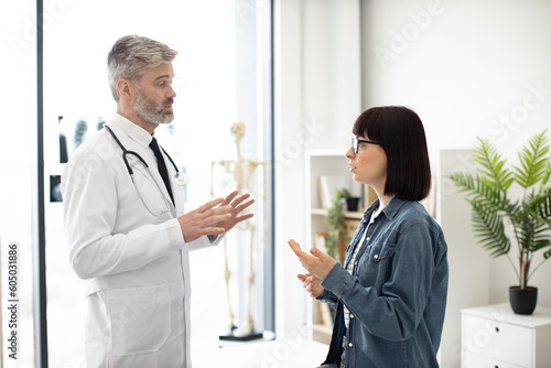 Attractive young woman holding hand on throat while describing health condition to bearded man in lab coat. Confident therapist giving consultation about illness to female patient at modern hospital.