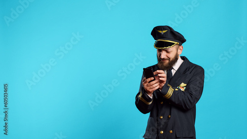 Aircrew captain scrolling through social media app, using smartphone with online internet in studio. Young airplane pilot playing with mobile phone and browser over blue background.