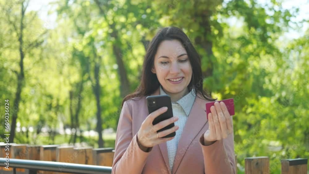 Caucasian young adult attractive woman in a suit smiles and makes a purchase via phone using a bank card Caucasian young adult attractive woman in a suit smiles and makes a purchase via phone using a bank card