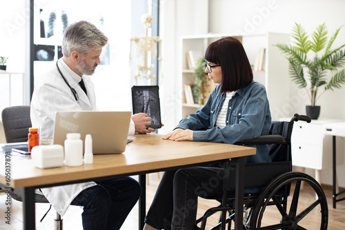 Confident adult in doctor's coat holding tablet while pretty lady in glasses sitting in wheelchair at writing desk in hospital. Efficient traumathologist analyzing CT scan image with patient at work.