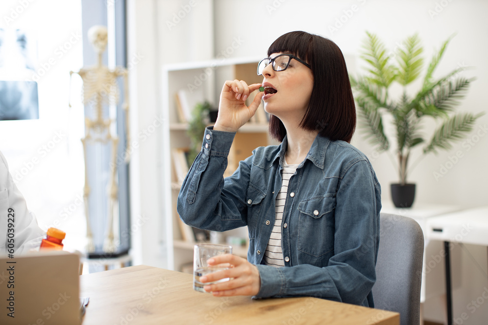 Attractive woman in everyday wear taking round pill with glass of water while sitting at office desk in modern office. Charming brunette in spectacles being prescribed antibiotics by family doctor. Attractive woman in everyday wear taking round pill with glass of water while sitting at office desk in modern office. Charming brunette in spectacles being prescribed antibiotics by family doctor.
