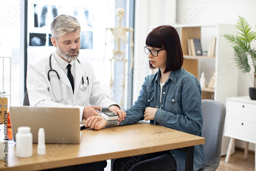 Confident bearded male with stethoscope turning on wearable tonometer applied on female wrist at doctor s office. Focused aged family physician measuring pulse rate of young caucasian woman.